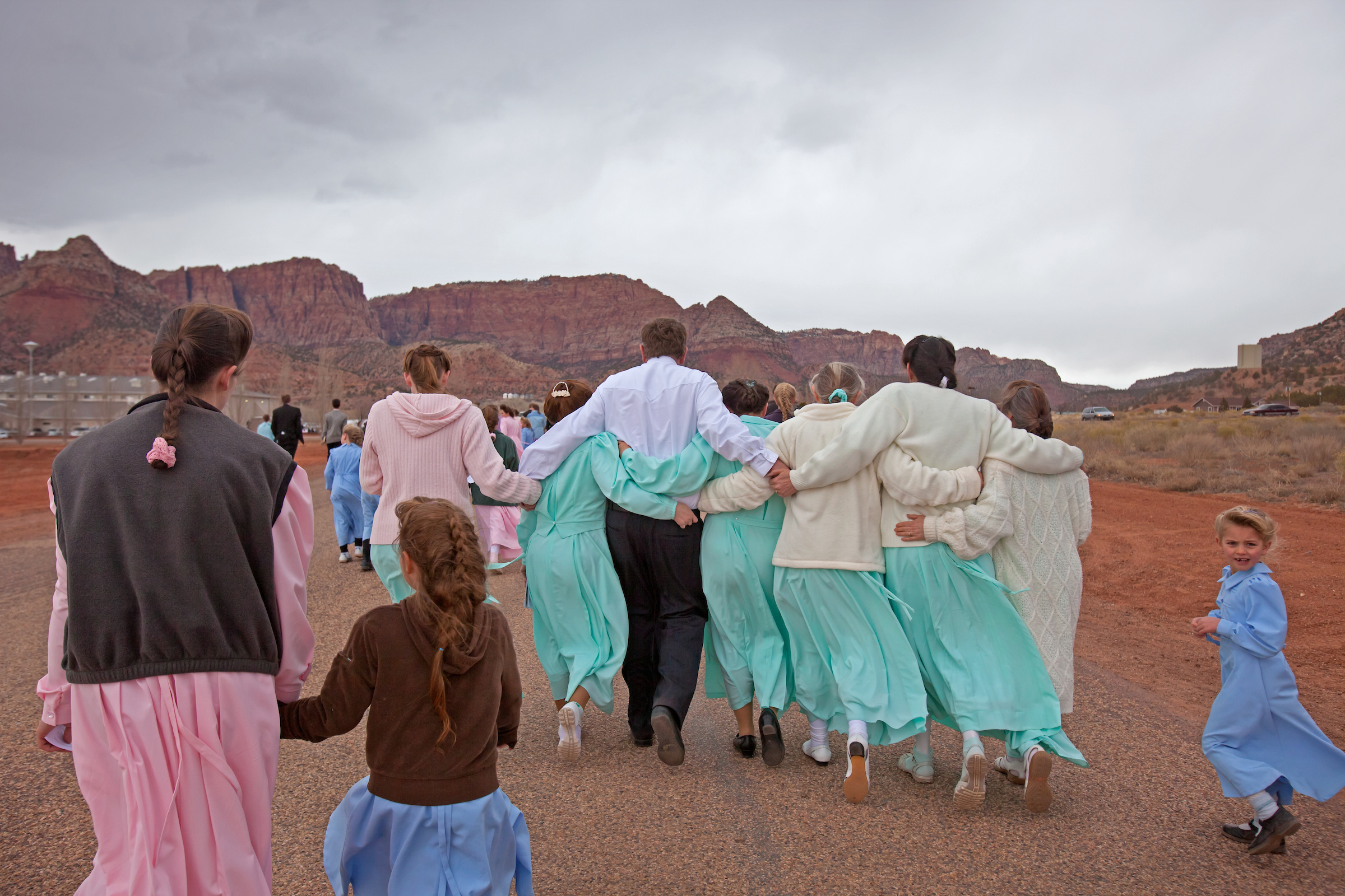 An FLDS man leaves the funeral service linking arms with six of his wives.